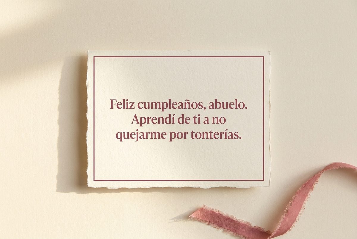Tarjeta de cumpleaños de papel crema hecho a mano, con borde deckle y un filete impreso en rosa oscuro, fotografiada en plano cenital. En la tarjeta se lee «Feliz cumpleaños, abuelo. Aprendí de ti a no quejarme por tonterías.». Al lado, una cinta de tela rosa deshilachada en la esquina inferior derecha.