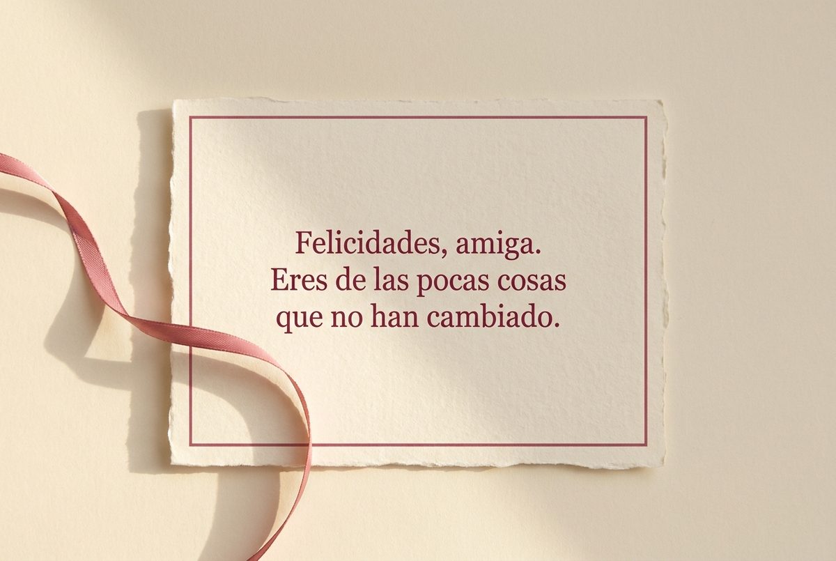 Tarjeta de cumpleaños de papel crema hecho a mano, con borde deckle y un filete impreso en rosa oscuro, fotografiada en plano cenital. En la tarjeta se lee «Felicidades, amiga. Eres de las pocas cosas que no han cambiado.». Al lado, una cinta de raso rosa que entra ondulando desde el borde izquierdo.