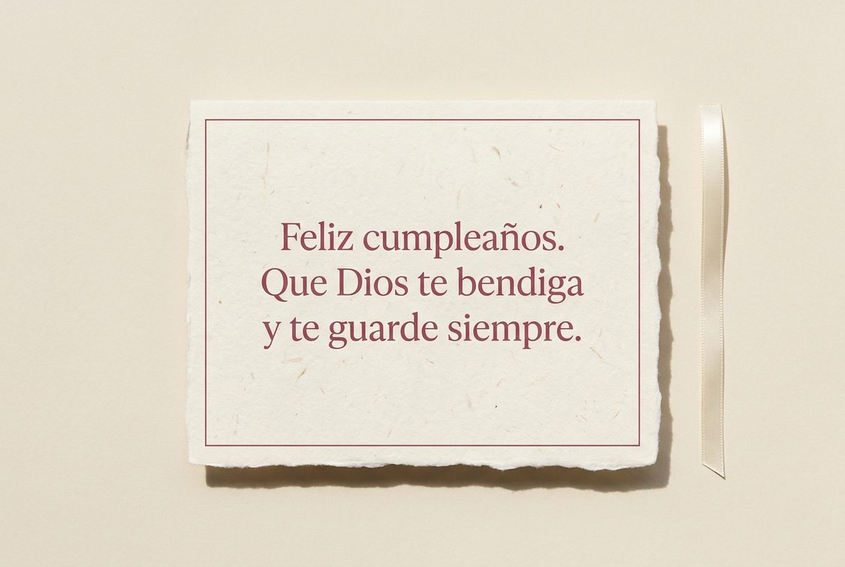 Tarjeta de cumpleaños de papel crema hecho a mano, con borde deckle y un filete impreso en rosa oscuro, fotografiada en plano cenital. En la tarjeta se lee «Feliz cumpleaños. Que Dios te bendiga y te guarde siempre.». Al lado, una cinta de raso crema estrecha junto al borde derecho.