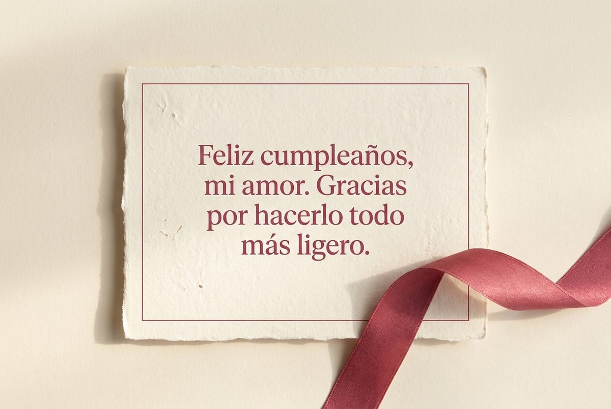 Tarjeta de cumpleaños de papel crema hecho a mano, con borde deckle y un filete impreso en rosa oscuro, fotografiada en plano cenital. En la tarjeta se lee «Feliz cumpleaños, mi amor. Gracias por hacerlo todo más ligero.». Al lado, una cinta de seda rosa intensa que se enrosca bajo la esquina inferior derecha.
