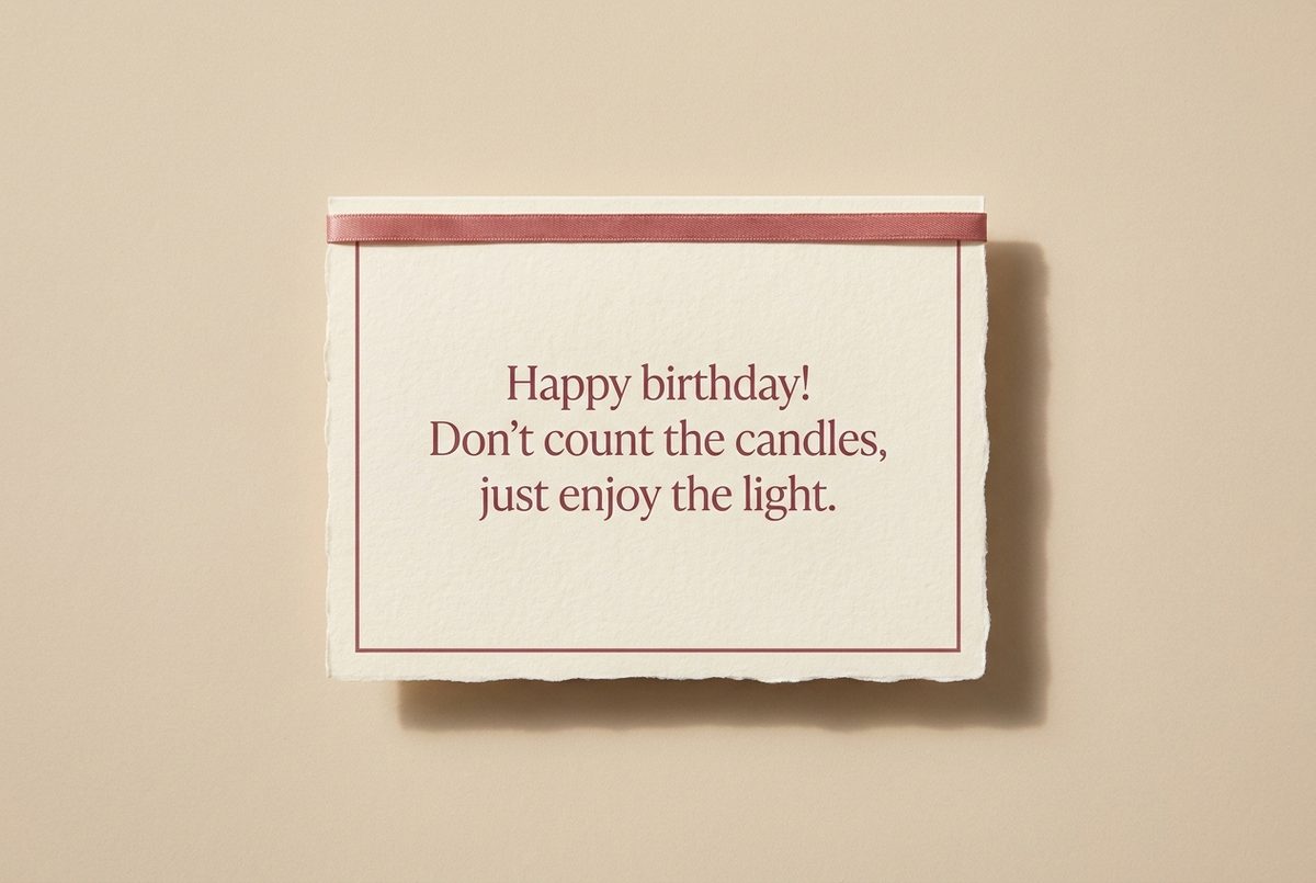 Tarjeta de cumpleaños de papel crema hecho a mano, con borde deckle y un filete impreso en rosa oscuro, fotografiada en plano cenital. En la tarjeta se lee «Happy birthday! Don't count the candles, just enjoy the light.». Al lado, una cinta rosa recta a lo largo del borde superior.
