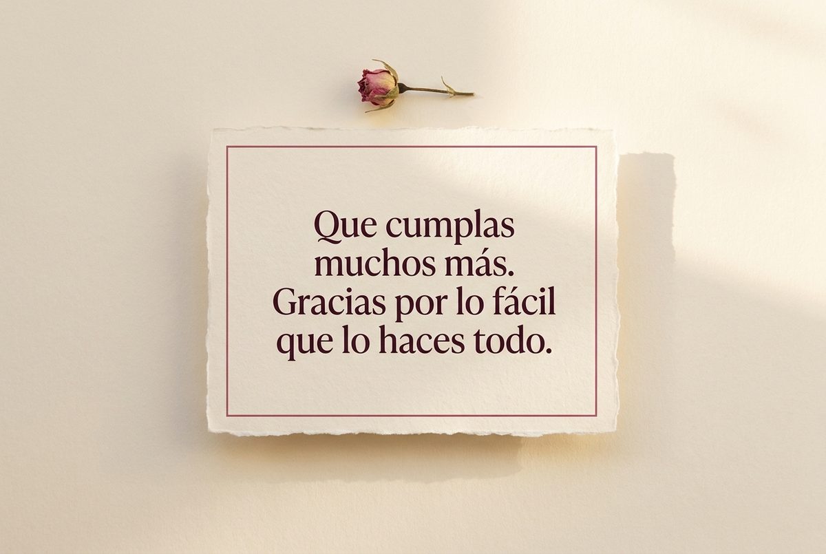 Tarjeta de cumpleaños de papel crema hecho a mano, con borde deckle y un filete impreso en rosa oscuro, fotografiada en plano cenital. En la tarjeta se lee «Que cumplas muchos más. Gracias por lo fácil que lo haces todo.». Al lado, un capullo de rosa seco colocado justo encima de la tarjeta.