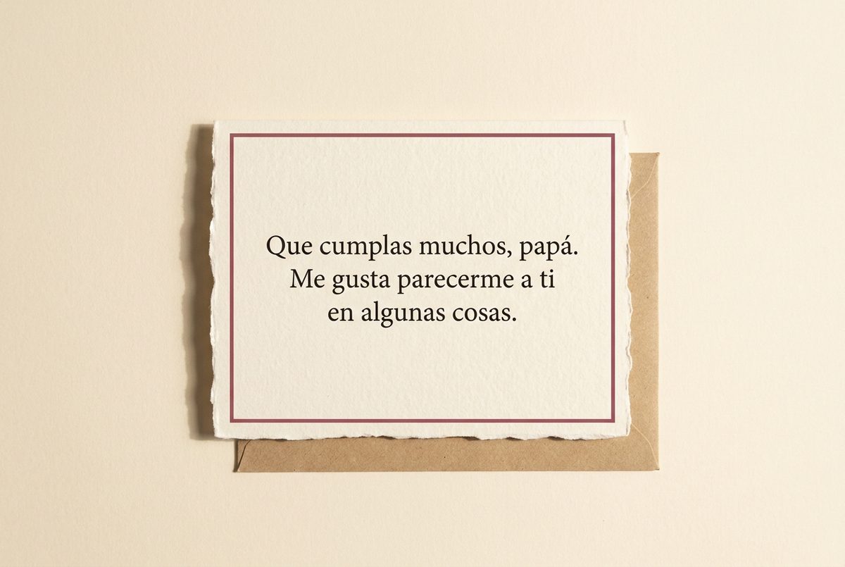 Tarjeta de cumpleaños de papel crema hecho a mano, con borde deckle y un filete impreso en rosa oscuro, fotografiada en plano cenital. En la tarjeta se lee «Que cumplas muchos, papá. Me gusta parecerme a ti en algunas cosas.». Al lado, un sobre de papel kraft bajo el borde inferior.