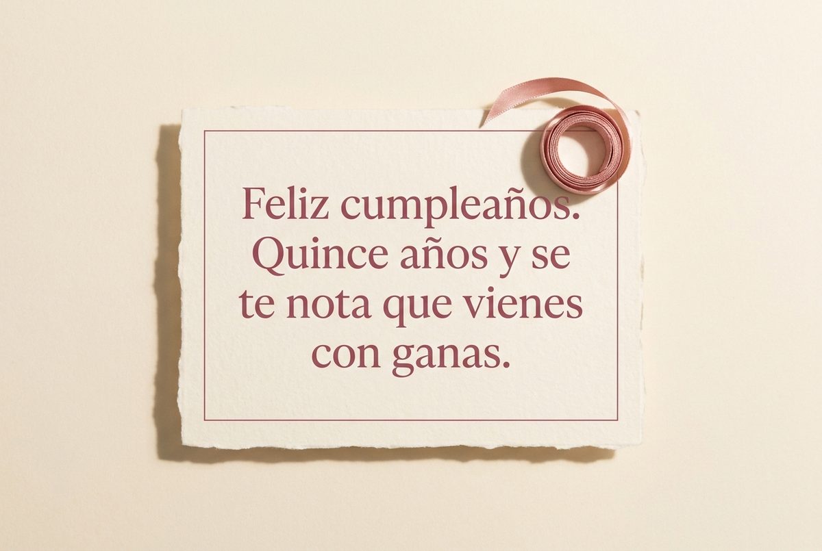 Tarjeta de cumpleaños de papel crema hecho a mano, con borde deckle y un filete impreso en rosa oscuro, fotografiada en plano cenital. En la tarjeta se lee «Feliz cumpleaños. Quince años y se te nota que vienes con ganas.». Al lado, un carrete de cinta rosa enrollado sobre la esquina superior derecha.