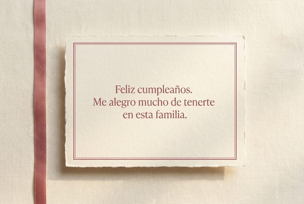 Tarjeta de cumpleaños de papel crema hecho a mano, con borde deckle y un filete impreso en rosa oscuro, fotografiada en plano cenital. En la tarjeta se lee «Feliz cumpleaños. Me alegro mucho de tenerte en esta familia.». Al lado, una cinta de raso rosa vertical a lo largo del borde izquierdo.